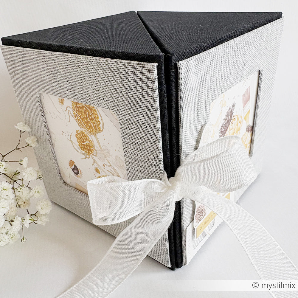 Graues und schwarzes Alexandra Renke PAPPKit Fotoalbum in Dreiecksform mit weißem Band, das neben kleinen weißen Blumen ausgestellt ist.
