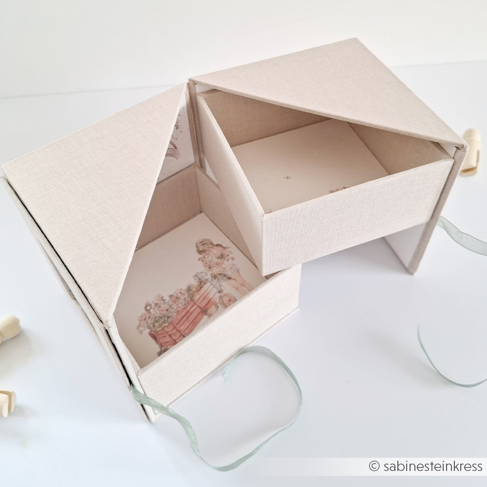 Eine cremefarbene, hausförmige Fotobox von Alexandra Renke lässt sich öffnen und gibt den Blick auf zwei Fächer frei, die mit Blumen- und Fahrradillustrationen verziert sind.