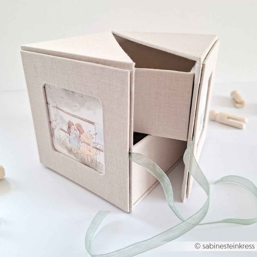 Eine cremefarbene Fotowürfelbox von Alexandra Renke, teilweise geöffnet mit einem Bändchen und einer Illustration von zwei Mädchen an der Seite.
