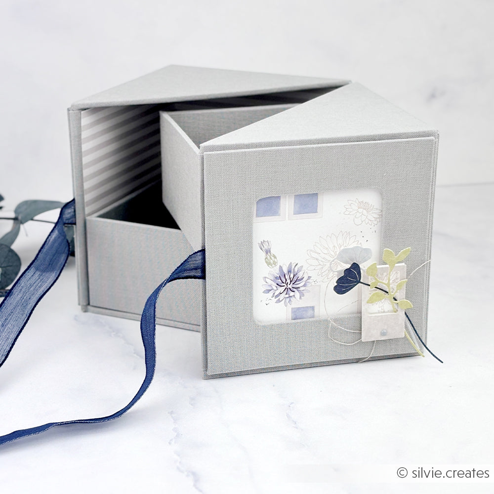 Die graue Alexandra Renke Fotobox mit blauer Schleife ist offen und zeigt im Inneren eine Blumenkarte und dekorative Blätter.