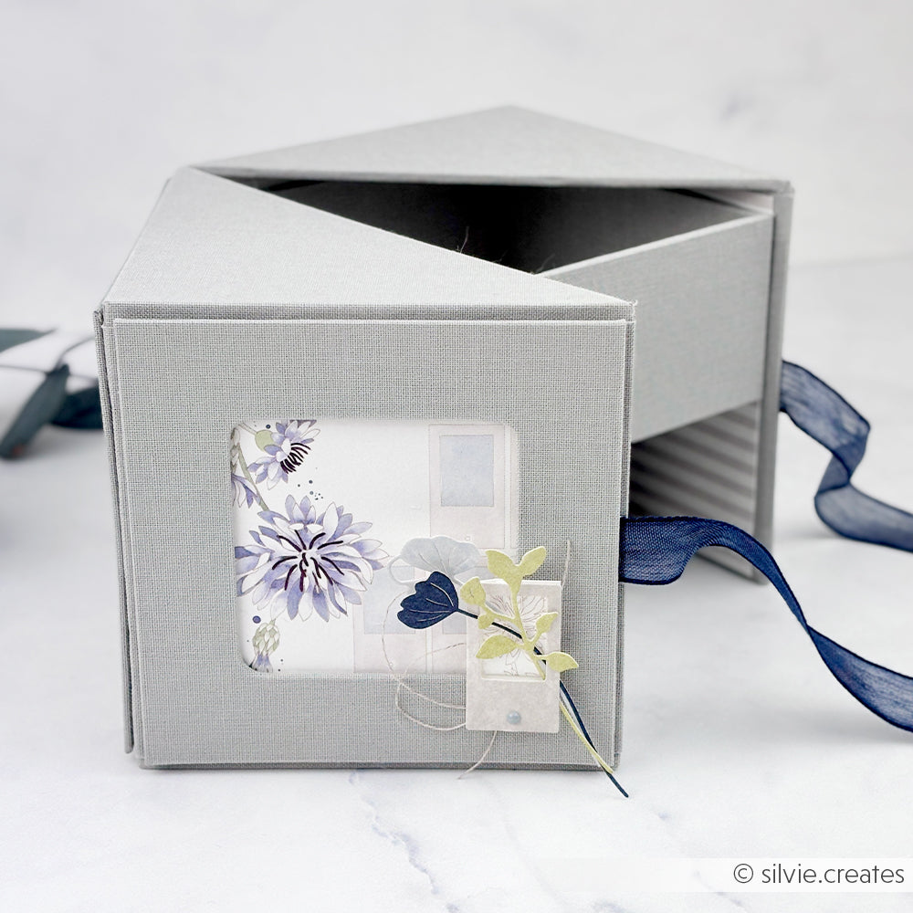 Eine graue sechseckige Alexandra Renke Fotowürfelbox mit Blumenkarte und Schleife, ausgestellt auf einer hellen Fläche.