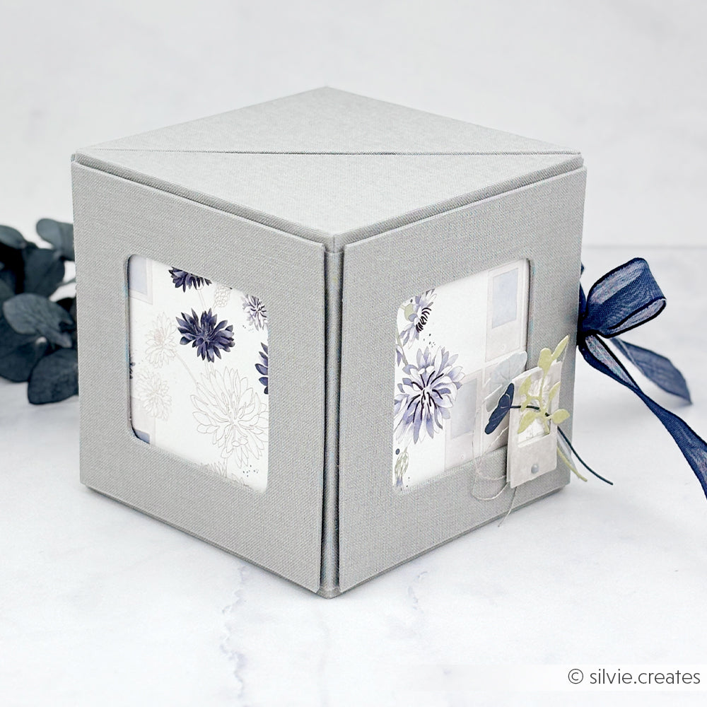 Eine mit grauem Stoff bezogene Alexandra Renke Fotowürfelbox mit floralem Interieur und blauem Band, ausgestellt auf einer weißen Fläche.
