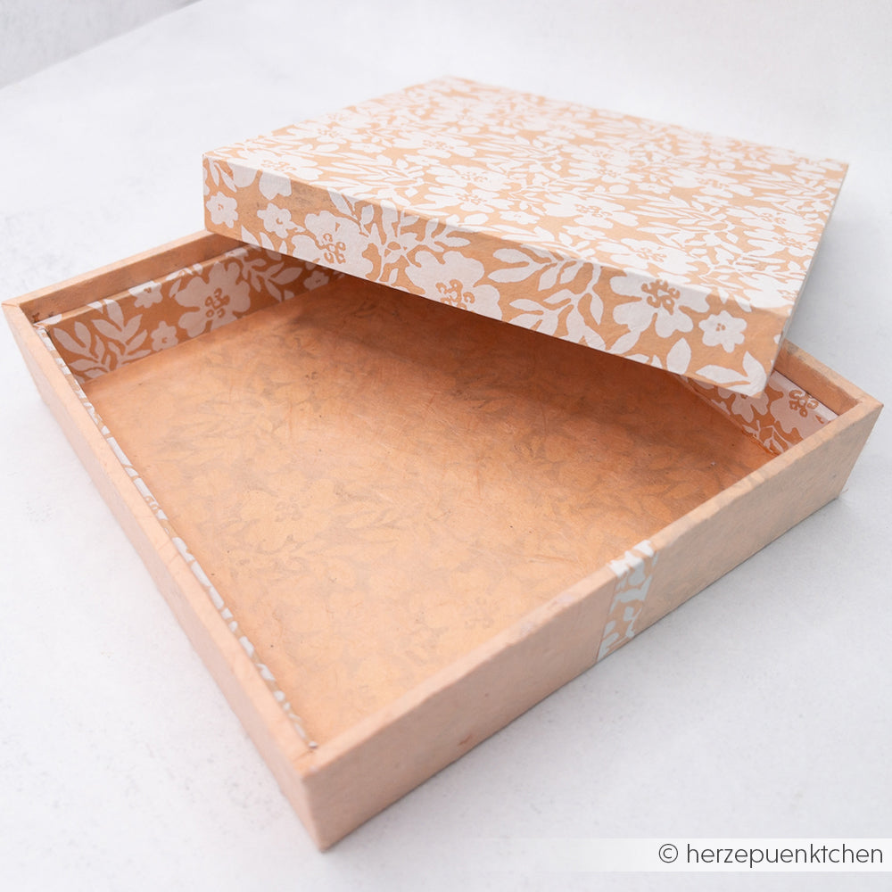 Eine dekorative, pfirsichfarbene Einsteckbox von Alexandra Renke mit weißem Blumenmuster, ideal zum Basteln von Geschenkverpackungen, steht teilweise geöffnet auf einer weißen Oberfläche.