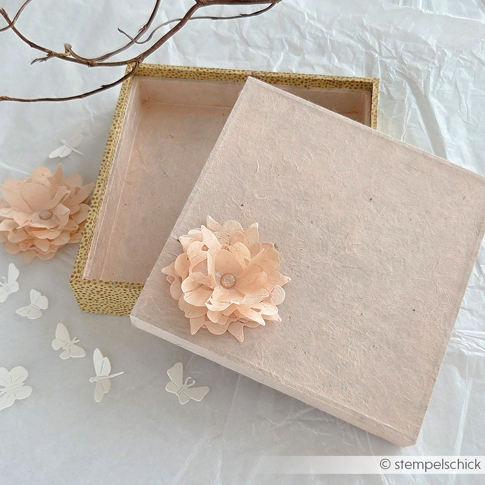 Quadratische Geschenkbox mit einer rosa Papierblume auf dem Deckel, umgeben von Blumen und Schmetterlingen - eine bezaubernde DIY-Geschenkverpackung mit dem PAPPKit von Alexandra Renke für Ihre besonderen Momente.