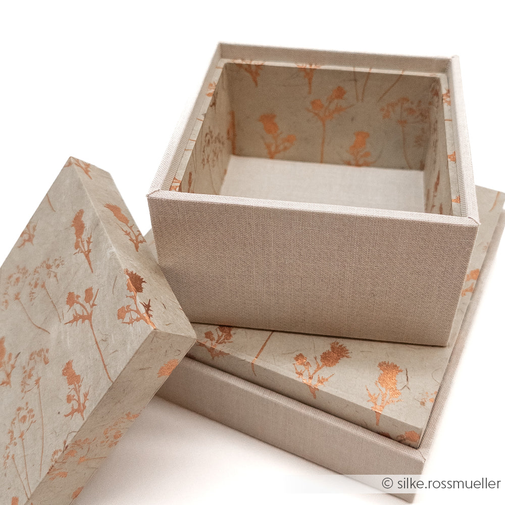 Zwei beigefarbene, floral gemusterte Alexandra Renke Geschenkverpackungen, eine offene und eine geschlossene, sind auf einer weißen Fläche gestapelt.