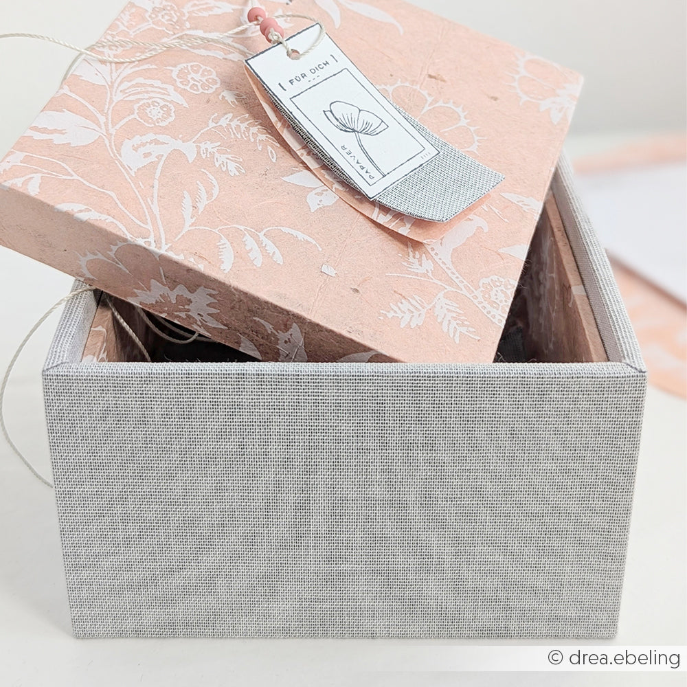 Eine graue und rosa geblümte Alexandra Renke Geschenkverpackung (PAPPKit Geschenkverpackungen) mit einem Anhänger, teilweise geöffnet auf einer hellen Oberfläche.