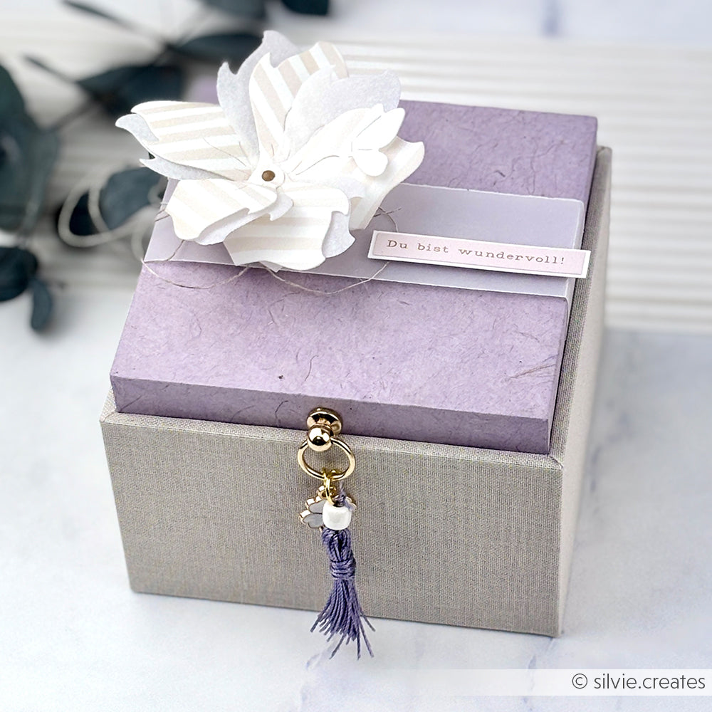 Eine lavendelfarbene und graue Alexandra Renke Geschenkbox mit einer weißen Blume und einem "Du bist wundervoll!"-Anhänger auf dem Deckel.