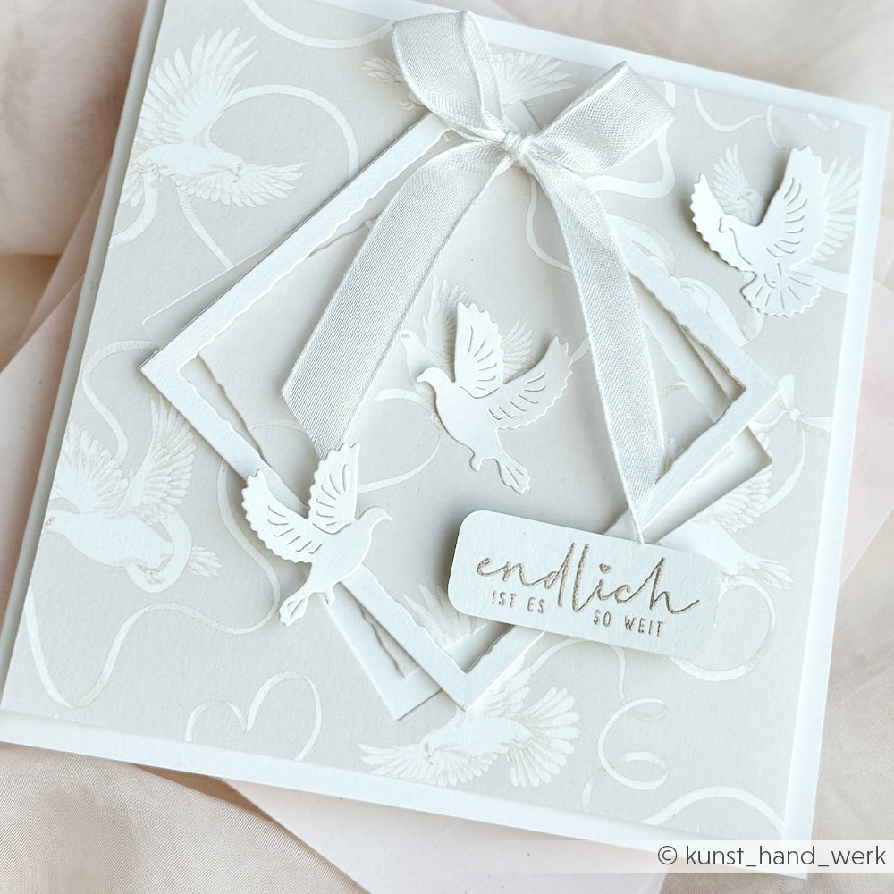 Weiße Karte von Alexandra Renke mit geprägten Tauben, Schleifenband, Lagenrahmen und der Aufschrift "Endlich ist es so weit". Perfekt für Papierprojekte - fügen Sie einfach Ihre Lieblings-Stempelkissen oder Holzstempel für eine persönliche Note hinzu.