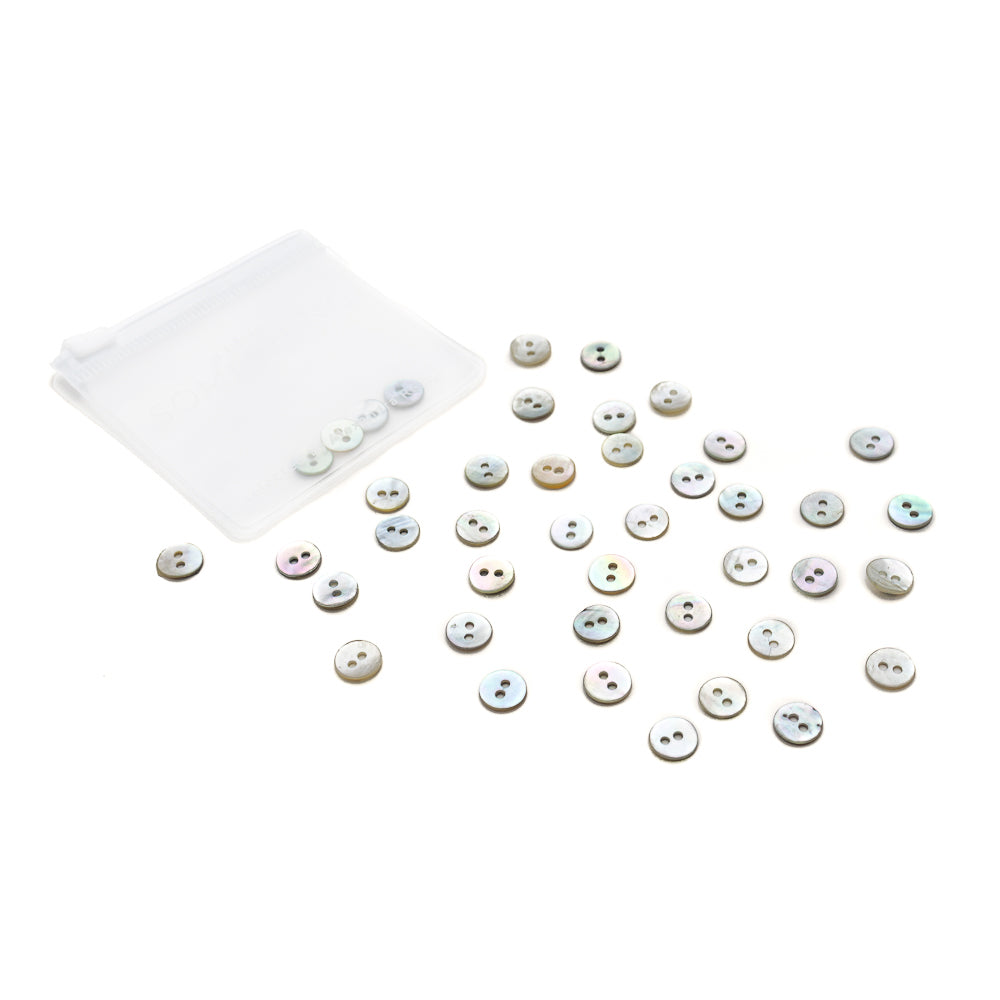 Kleine weiße Muschelknöpfe von Alexandra Renke verstreut neben einem durchsichtigen Plastikbeutel auf weißem Hintergrund.