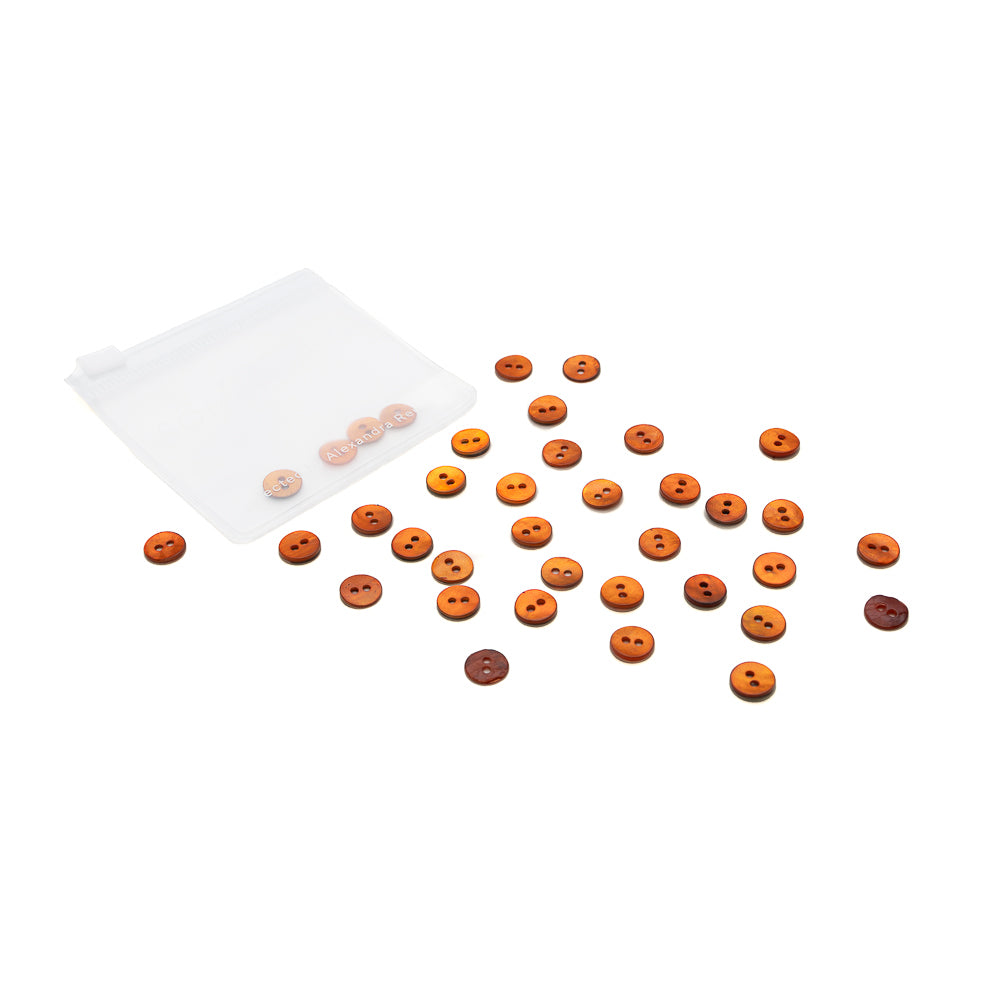Ein durchsichtiges Täschchen von Alexandra Renke enthält verstreute braune und orangefarbene Muschelknöpfe in verschiedenen Schattierungen, deren Spiegelglanz vor weißem Hintergrund das Licht auffängt.