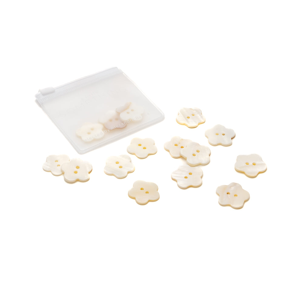 Ein Set kleiner, schimmernder Akoyaknöpfe in Blumenform von Alexandra Renke und ein durchsichtiges Reißverschlussetui werden auf weißem Hintergrund präsentiert.