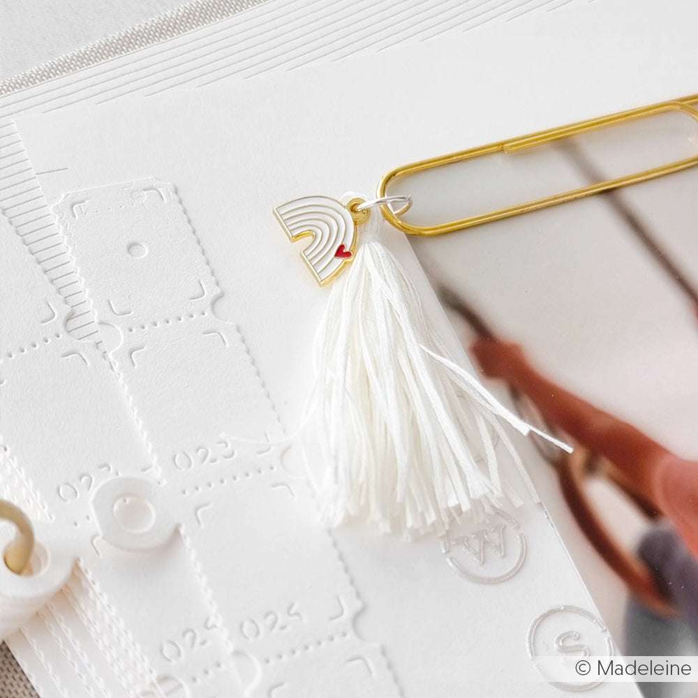 Goldfarbene Büroklammer mit weißer Quaste und kleinem Regenbogen-Anhänger von Alexandra Renke, angeklippt an weißes Strukturpapier - ideal für Buchbindearbeiten oder ausgefallene Papierprojekte.