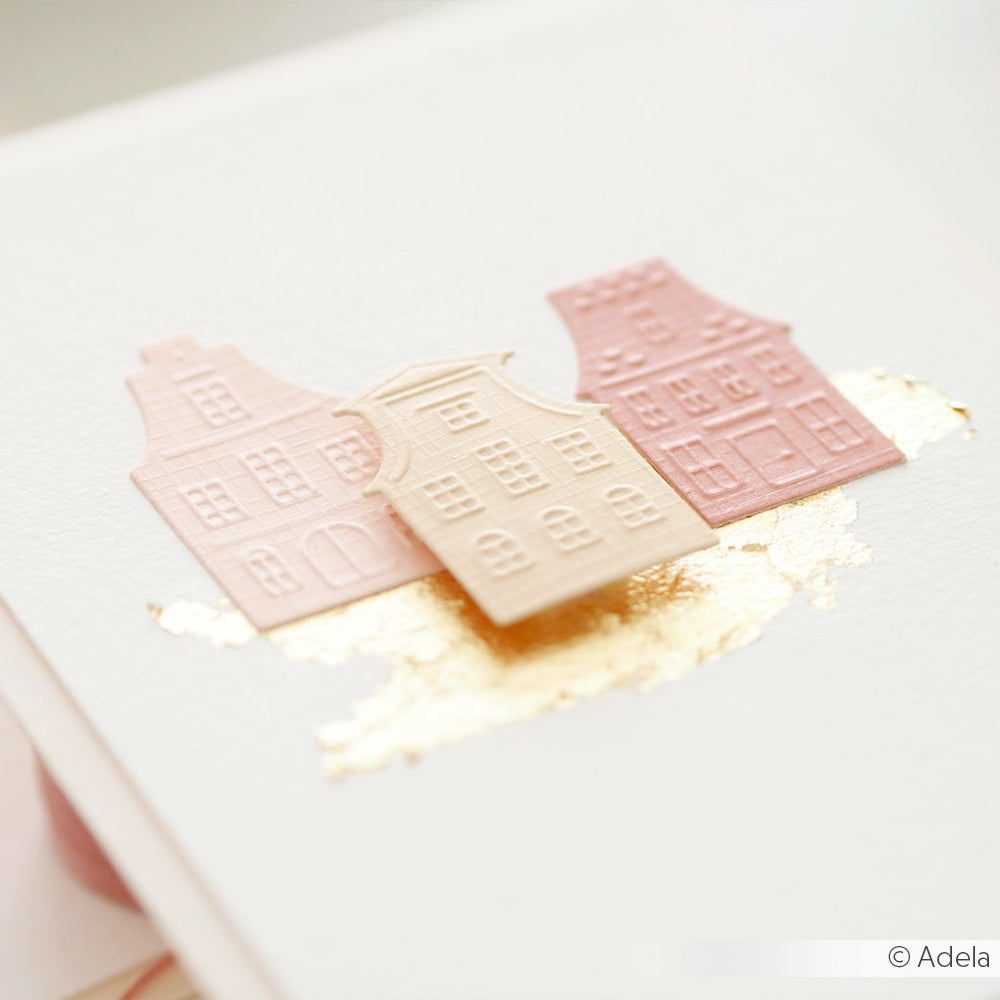 Drei pastellfarbene Papierhäuser, die mit dem Schneide- und Prägefolder Kleine Stadthäuser Stanzsysteme von Alexandra Renke gestaltet wurden, sitzen auf einer weißen Karte, die im Hintergrund mit Goldfolie akzentuiert ist.