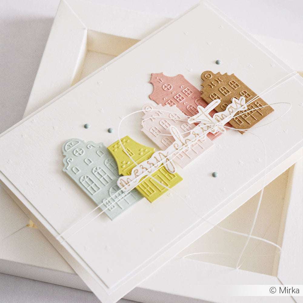 Eine handgefertigte Weihnachtskarte von Alexandra Renke mit pastellfarbenen Kleinen Stadthäusern und dem Schriftzug "Frohe Weihnachten" auf weißem Cardstock, hergestellt mit Stanzsystemen und einem Cut & Emboss Folder für zusätzliche Details.