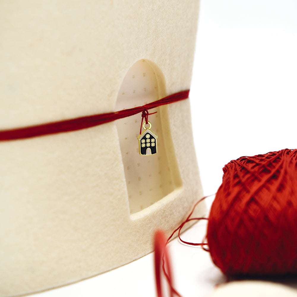 Eine rote Kordel von Alexandra Renke mit einem Schmuckanhänger und Charm Haus schwarz wird um einen cremefarbenen Gegenstand gebunden, daneben ein Knäuel roter Faden - ideal für Buchbindearbeiten oder um Ihren Bastelarbeiten einen einzigartigen Akzent zu verleihen.