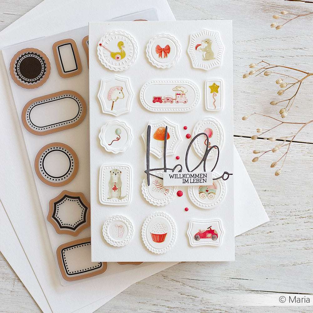 Grußkarte von Alexandra Renke mit geprägten Baby- und Spielzeugaufklebern im Vintage-Stil, die mit Cut & Emboss Folder-Techniken hergestellt wurden, ausgestellt auf einem Holztisch.