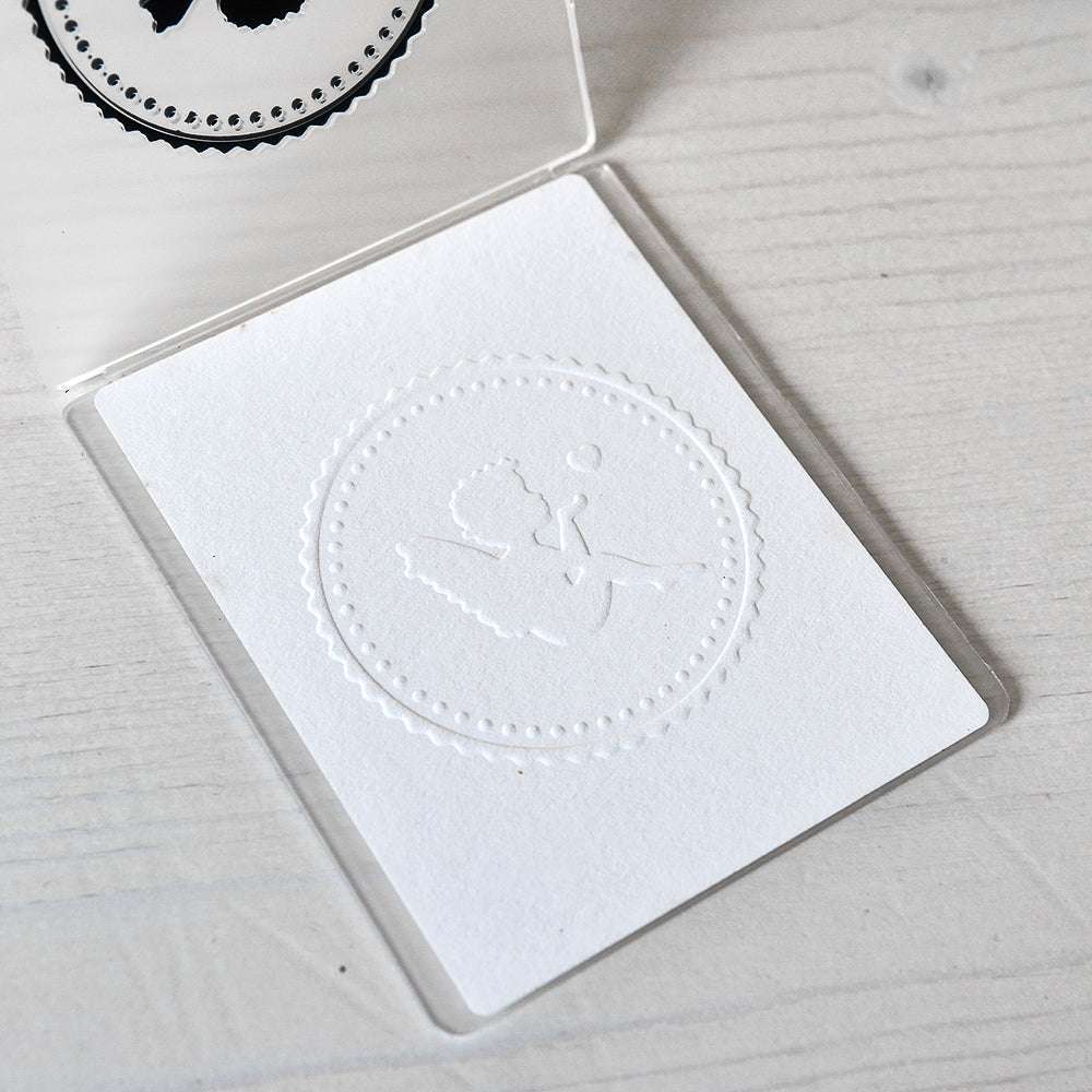 Weiße Karte mit einem geprägten Engel mit Herz, der eine Schleife in einem gepunkteten Kreis hält, erstellt mit einem Alexandra Renke Prägefolder, auf einer hellen Holzoberfläche.