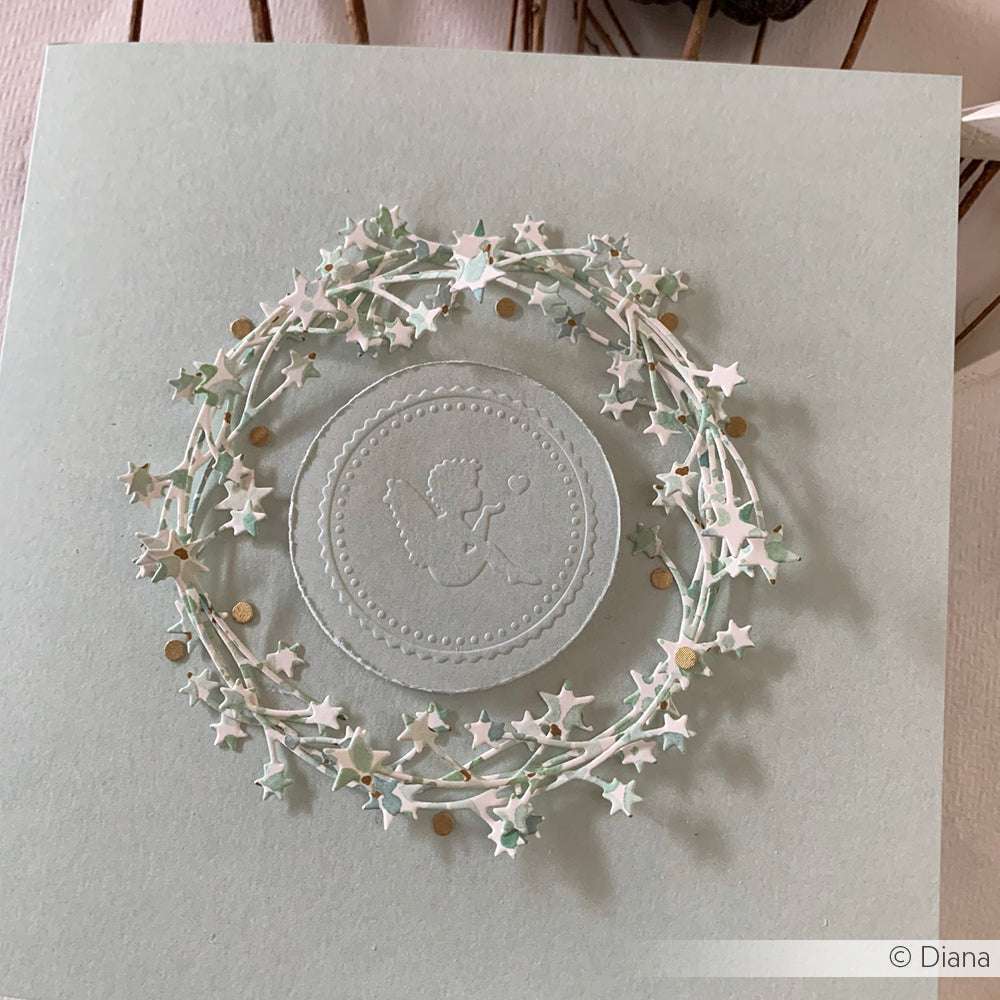 Ein kreisförmiger Kranz aus weißen Sternen umgibt einen geprägten Engel mit Herz auf einer hellgrauen Karte, die mit dem Alexandra Renke Papier prägen Embossing Folder hergestellt wurde.