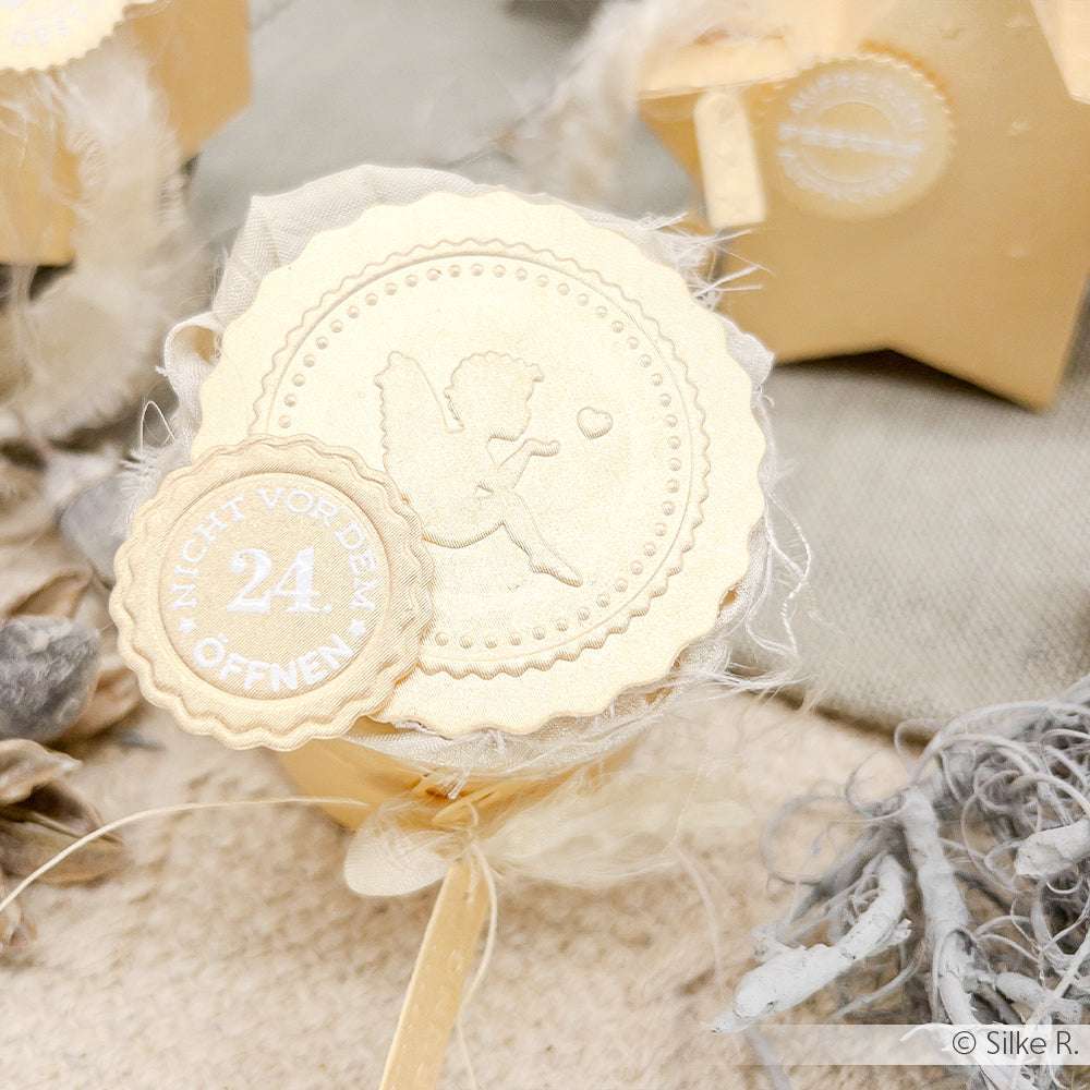 Runde goldene Geschenkbox von Alexandra Renke mit einem "Engel mit Herz" und "24. NICHT VOR DEM ÖFFNEN" auf dem Deckel, Papier geprägt wie von einer Prägemappe, umgeben von festlichem Dekor.