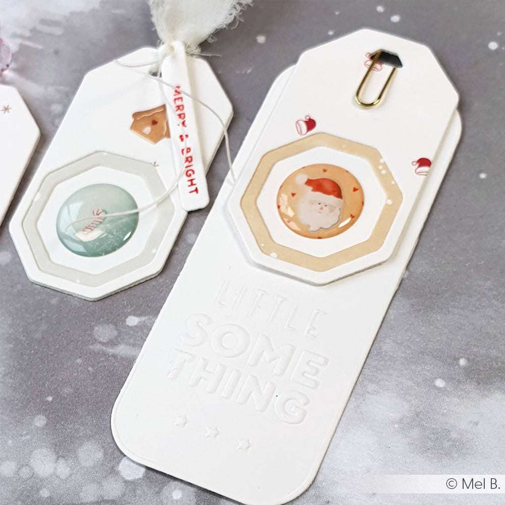 Zwei handgefertigte Geschenkanhänger von Alexandra Renke mit Lagenpapierdesigns, die mit Stanzsystemen hergestellt wurden - einer zeigt den Weihnachtsmann, der andere eine festliche Szene - perfekt, um Ihren Geschenken eine besondere Note zu verleihen.