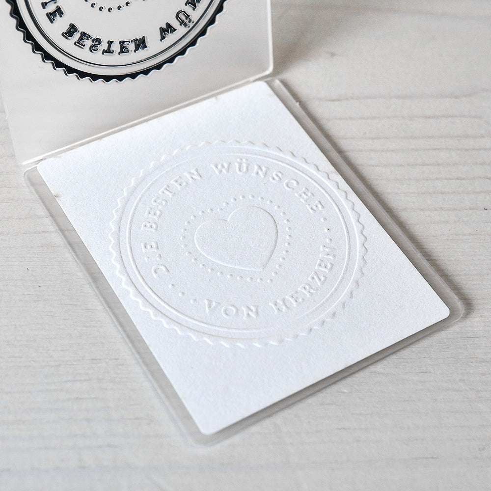 Weiße Karte mit geprägtem Herz und deutschem Text "DIE BESTEN WÜNSCHE VON HERZEN" auf einer hellen Holzoberfläche, hergestellt mit einem Alexandra Renke Prägefolder für elegantes Papier prägen.