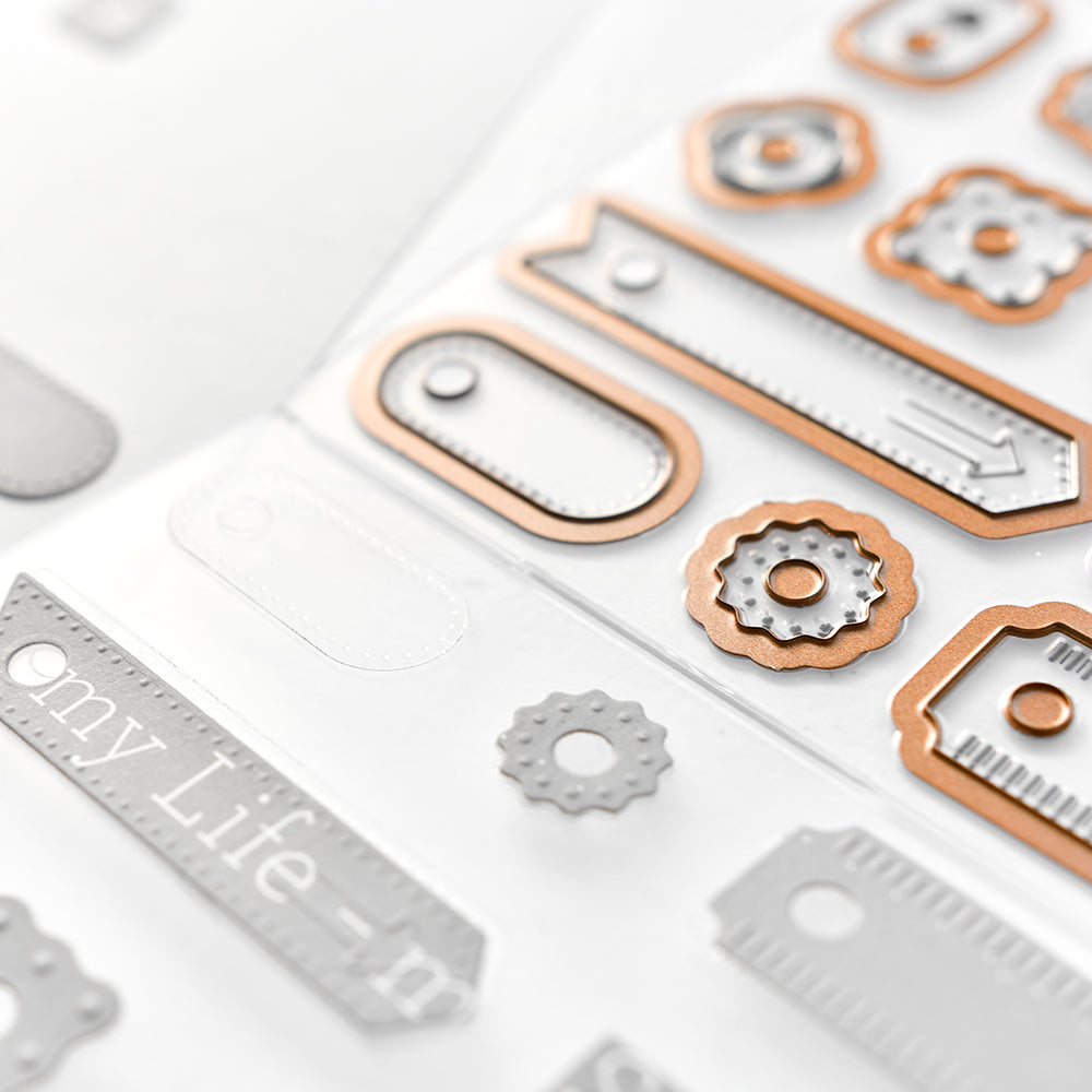 Nahaufnahme von verschiedenen metallischen und weißen Aufklebern in Form von Etiketten und Zahnrädern auf einer durchsichtigen Plastikfolie von Alexandra Renke, perfekt für Verstärker Formen oder die Verwendung mit Cut & Emboss Folder Stanzsystemen.