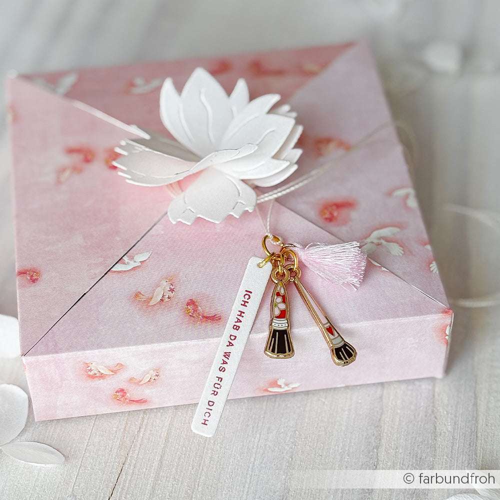 Eine rosafarbene, geblümte Geschenkbox von Alexandra Renke mit einem weißen Lotus, Schlüsselanhänger und einem Charms Pinsel Trio - ideal für Buchbindearbeiten oder zum sicheren Aufbewahren Ihrer Schmuckanhänger.