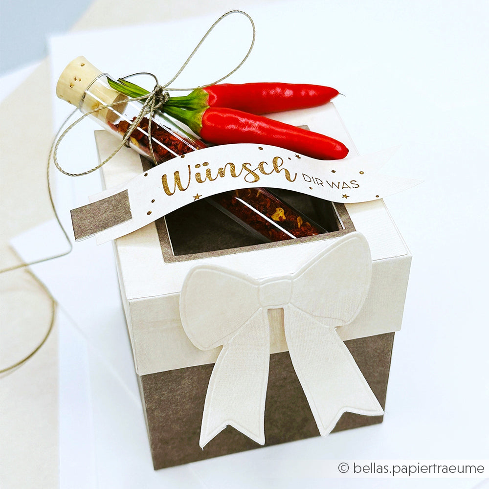 Geschenkbox von Alexandra Renke, verziert mit einer weißen Schleife, zwei roten Chilischoten, einem Glasröhrchen und einem Anhänger mit der Aufschrift "Wünsch dir was".