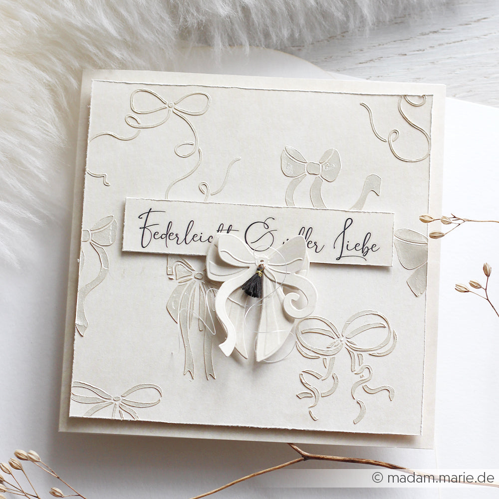 Elegante cremefarbene Karte, die mit dem Schneide- und Prägefolder von Alexandra Renke hergestellt wurde, mit geprägten Schleifen, einer Schleifenpracht in der Mitte und einem Text in deutscher Kalligraphie.