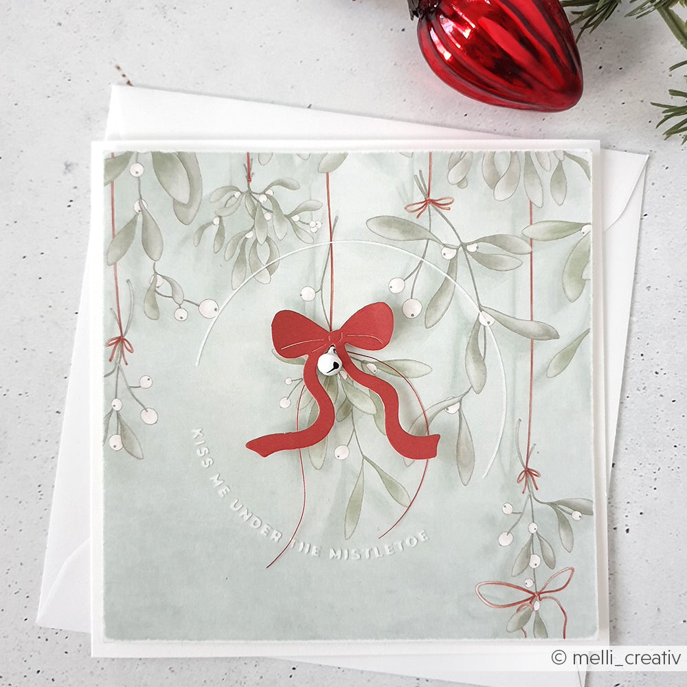 Quadratische Karte mit Mistelzweig, rotem Band in Schleifenpracht und "KISS ME UNDER THE MISTLETOE" auf hellem Hintergrund - gestaltet mit Alexandra Renke Cut & Emboss Folder Stanzsysteme für elegante Details.