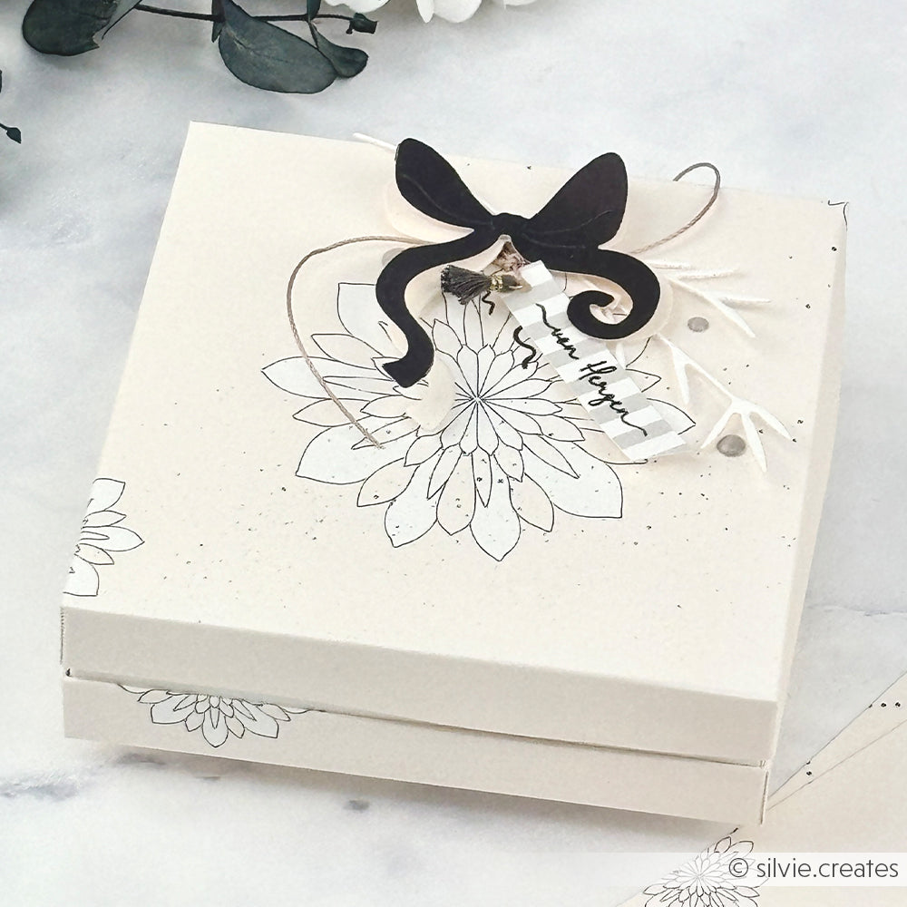 Weiße Geschenkschachtel von Alexandra Renke mit floralen Linien, schwarzer Schleife, kleinem Stern und dem Anhänger "Beste Wünsche" - ideal, um Ihre Schleifenpracht zu präsentieren oder Cut & Emboss Folder-Projekte zu verschönern.