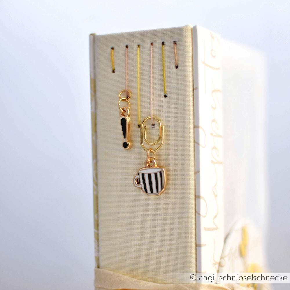 Drei goldene Lesezeichen von Alexandra Renke - eine Büroklammer, ein Ausrufezeichen und eine gestreifte Kaffeetasse - hängen an einem Buchrücken und verleihen Ihren Buchbindearbeiten und Papierprojekten Stil.