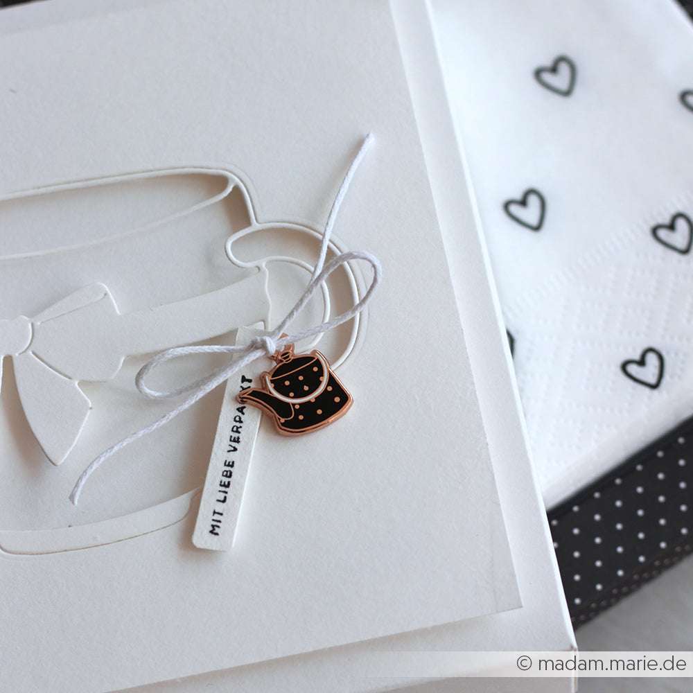 Weiße Grußkarte von Alexandra Renke mit Glasmotiv, Schleife und kleinem schwarzen Teekannenanhänger - ideal für Schmuck selber machen oder Charms Kaffeezeit - neben Servietten mit Herzmotiv.