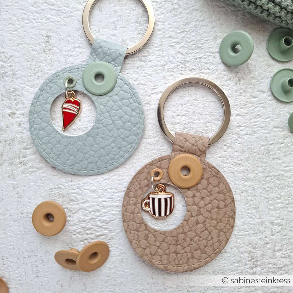 Zwei runde Leder-Schlüsselanhänger von Alexandra Renke mit Buchbindearbeiten und Kaffeezeit-Anhängern - ein Herz und eine Kaffeetasse -, die von Druckknöpfen umgeben sind.