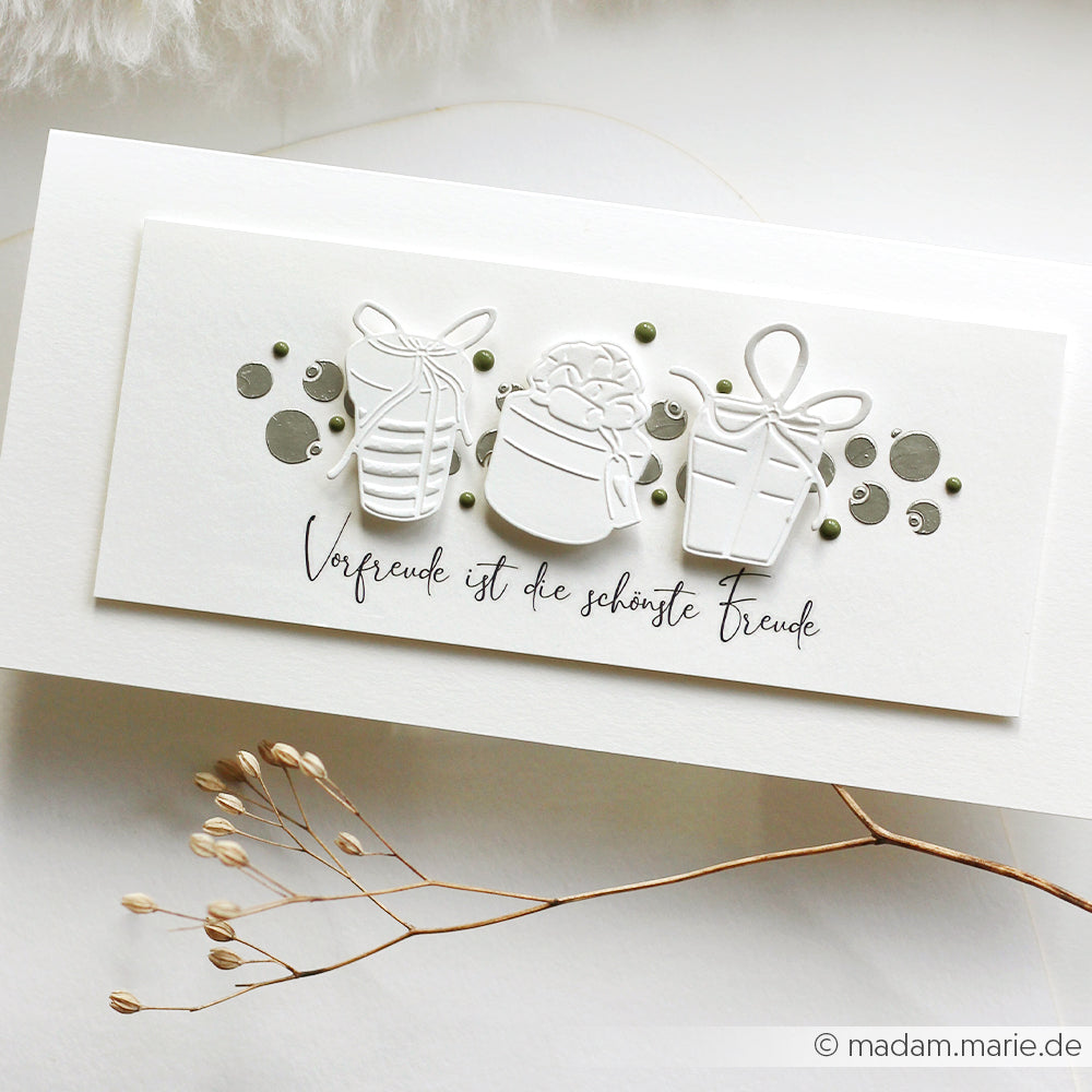 Eine von Alexandra Renke handgefertigte Freudenpäckchen-Karte zeigt geprägte weiße Geschenkschachteln, die mit einem Cut & Emboss Folder hergestellt wurden, deutschen Text, grüne Punkte und getrocknetes Gras.
