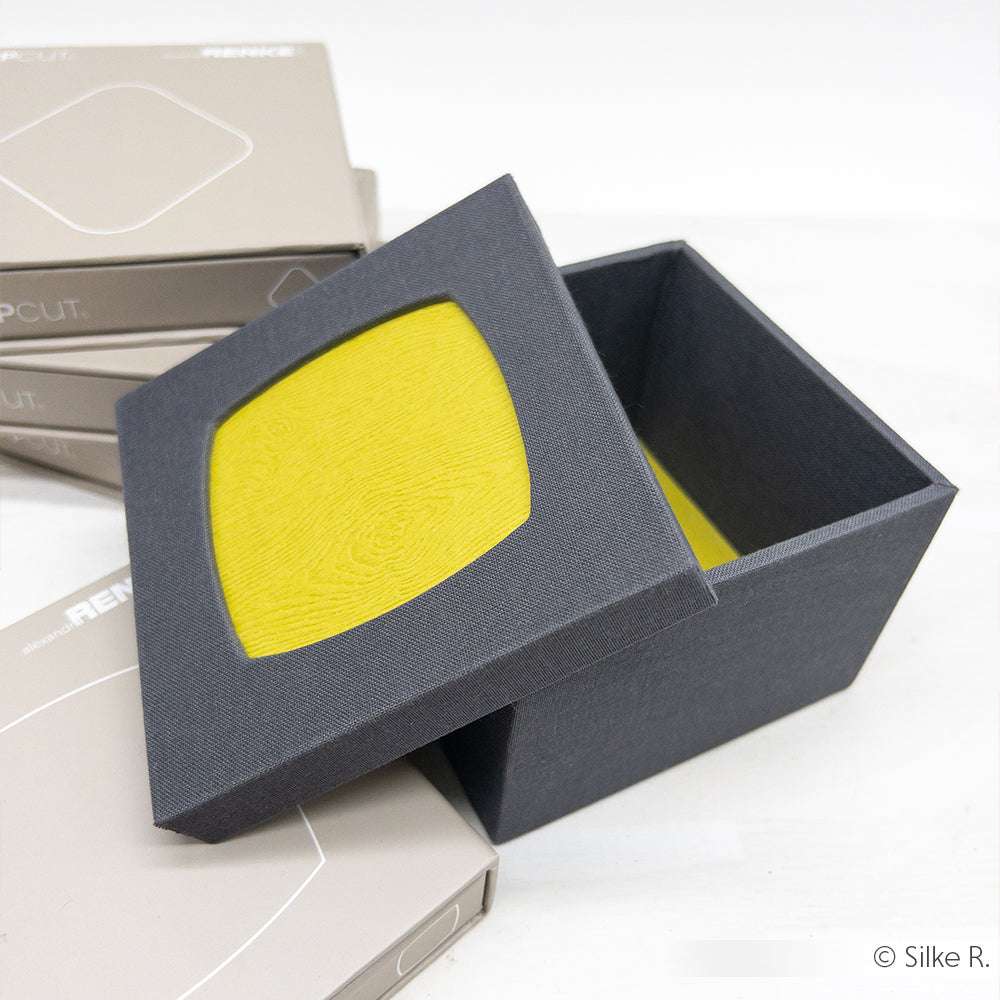 Eine schwarze, quadratische Alexandra Renke-Geschenkbox mit gelbem Innenleben und teilweise abnehmbarem Deckel, hergestellt mit der PAPPcut®-Technologie, steht neben gestapelten beigen Boxen - perfekt zum Buchbinden oder Präsentieren feiner Gegenstände.