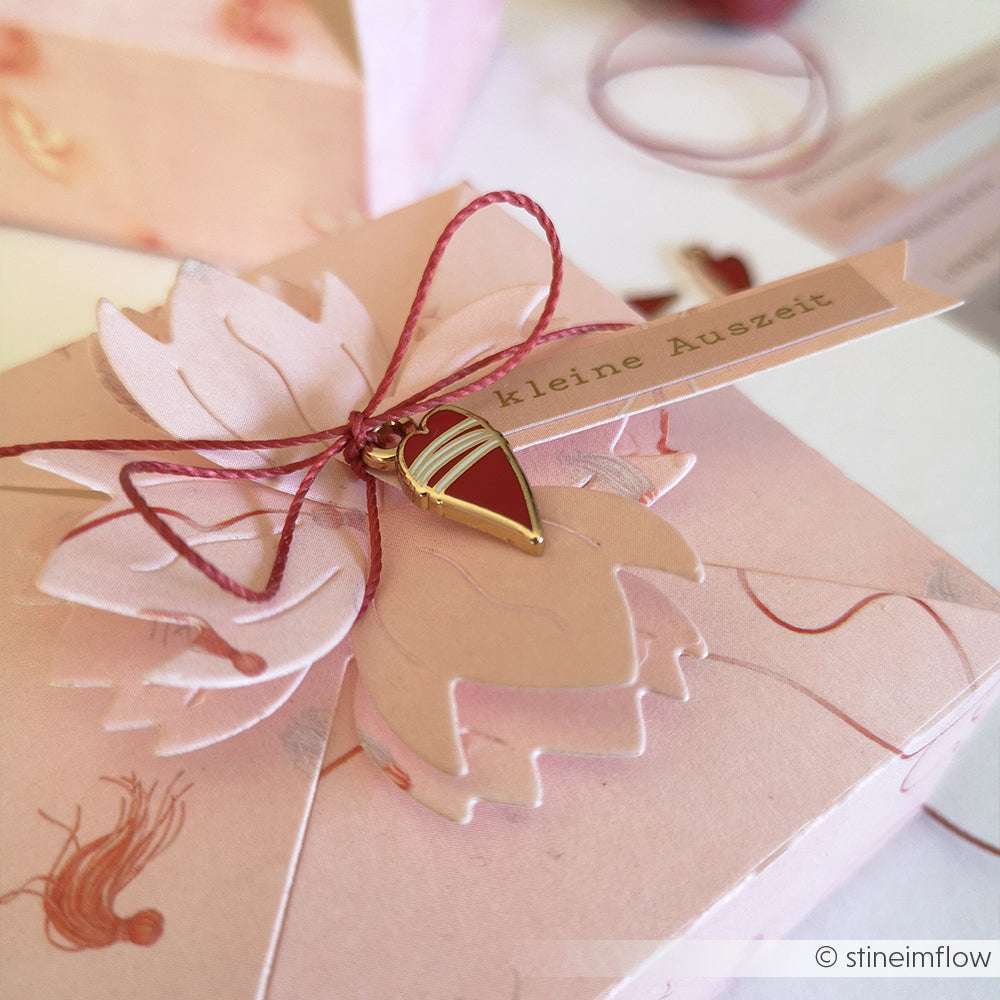 Eine rosafarbene Alexandra Renke Geschenkbox mit einem blumenförmigen Papier, Charms Herzen Anhänger und einem mit roter Kordel gebundenen "kleine Auszeit"-Anhänger - ideal für Buchbindearbeiten oder zur Aufbewahrung besonderer Andenken.