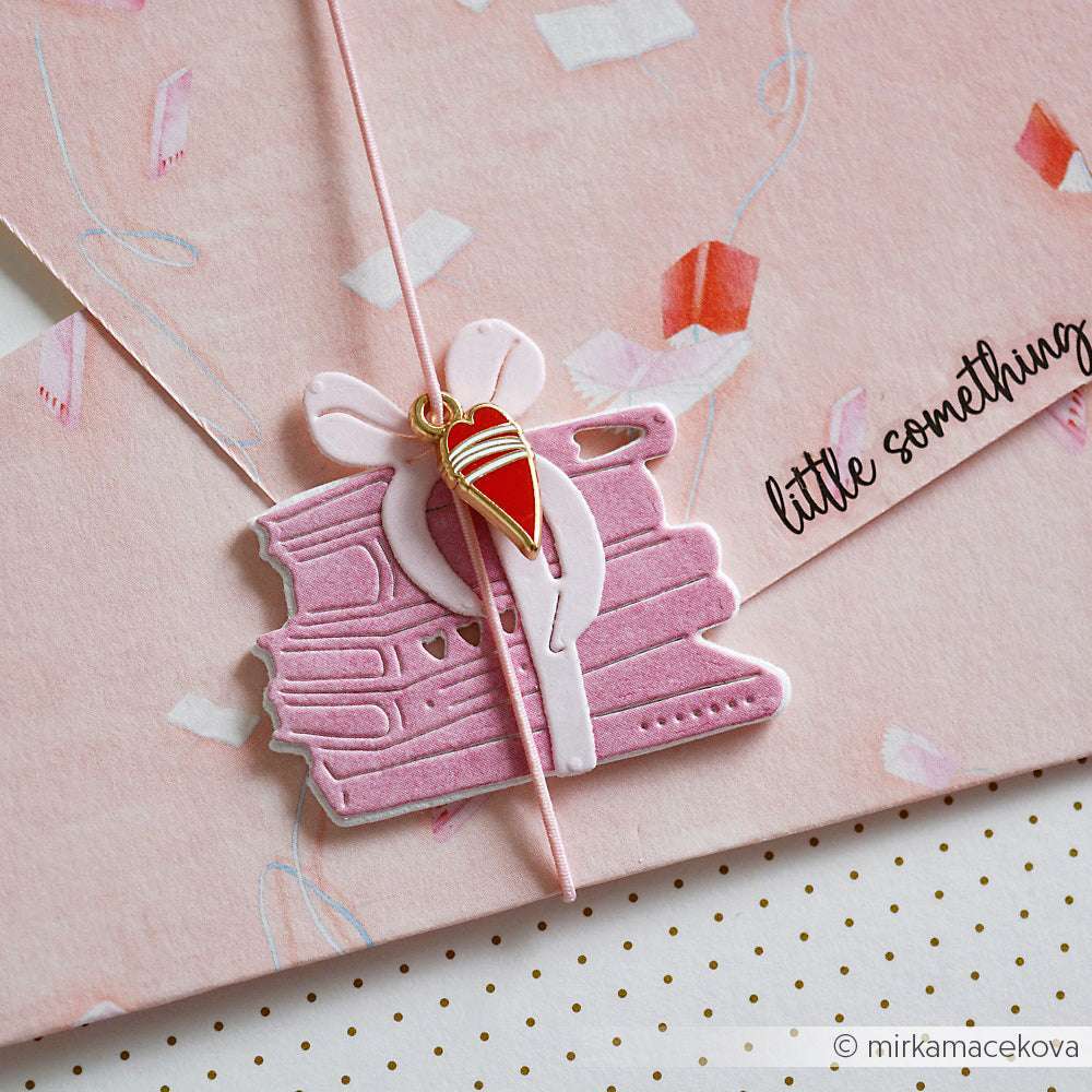 Ein Stapel rosafarbener Bücher, mit Schleife gebunden, und ein Alexandra Renke Charms Herzen Trio Herzanhänger ruhen auf einem rosafarbenen Umschlag mit der Aufschrift "Kleinigkeit" - das perfekte Bastelzubehör oder Schmuckanhänger für besondere Geschenke.