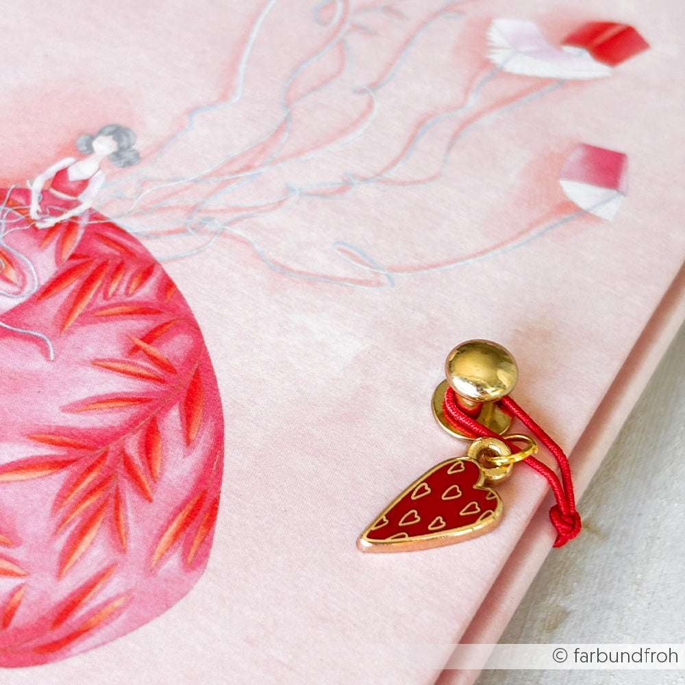 Ein goldener und roter Schmuckanhänger in Herzform von Alexandra Renke mit roter Kordel ruht auf einem rosa illustrierten Notizbuch - ideal für Buchbindearbeiten und perfekt für Ihre Charms Herzen Trio Sammlung.