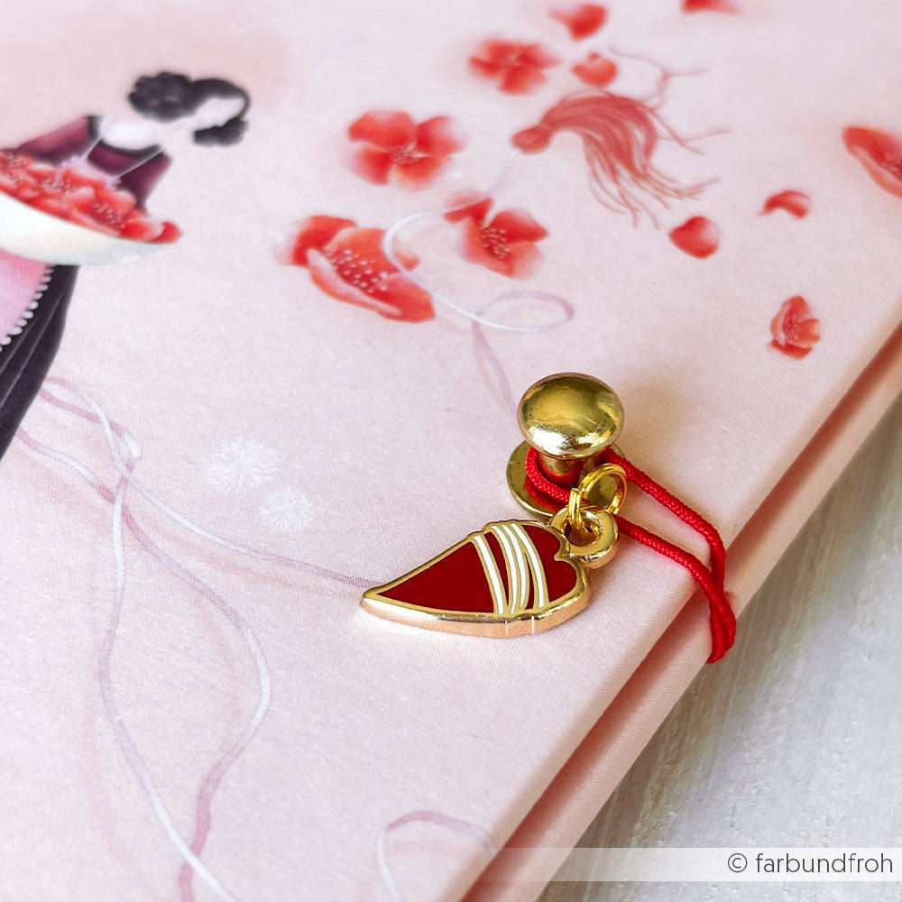 Alexandra Renke roter Schmuckanhänger in Herzform mit goldenen Akzenten an einer roten Schnur, ausgestellt auf einem rosa geblümten Notizbuch - ein bezauberndes Accessoire für jede Buchbindezubehör-Kollektion.