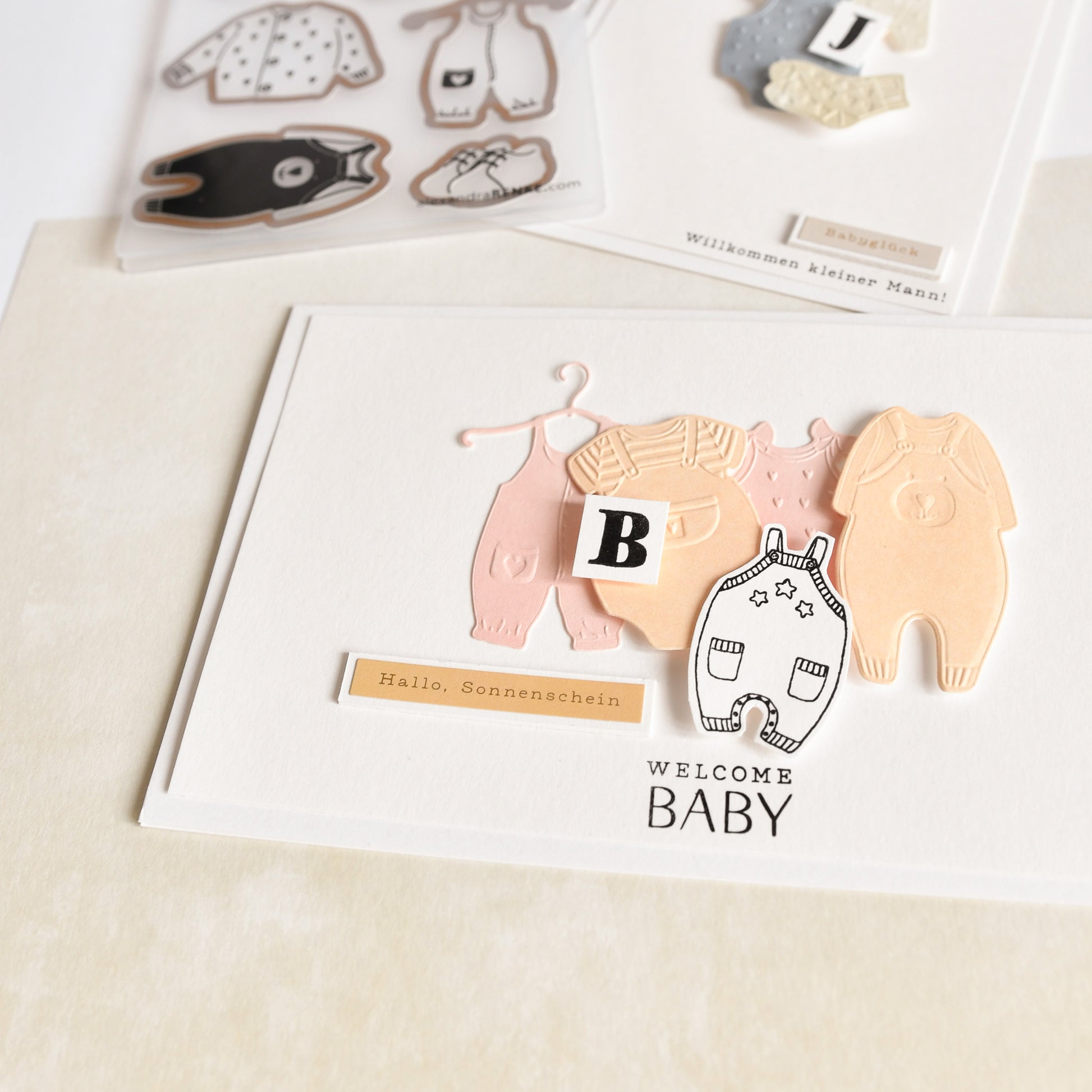 Grußkarte mit Alexandra Renke Designpapieren, ausgeschnittener Babykleidung, einem Block mit dem Buchstaben B und dem Text "Welcome Baby" auf hellem Hintergrund - wunderschön gestaltet mit unserem Cut & Emboss Folder.