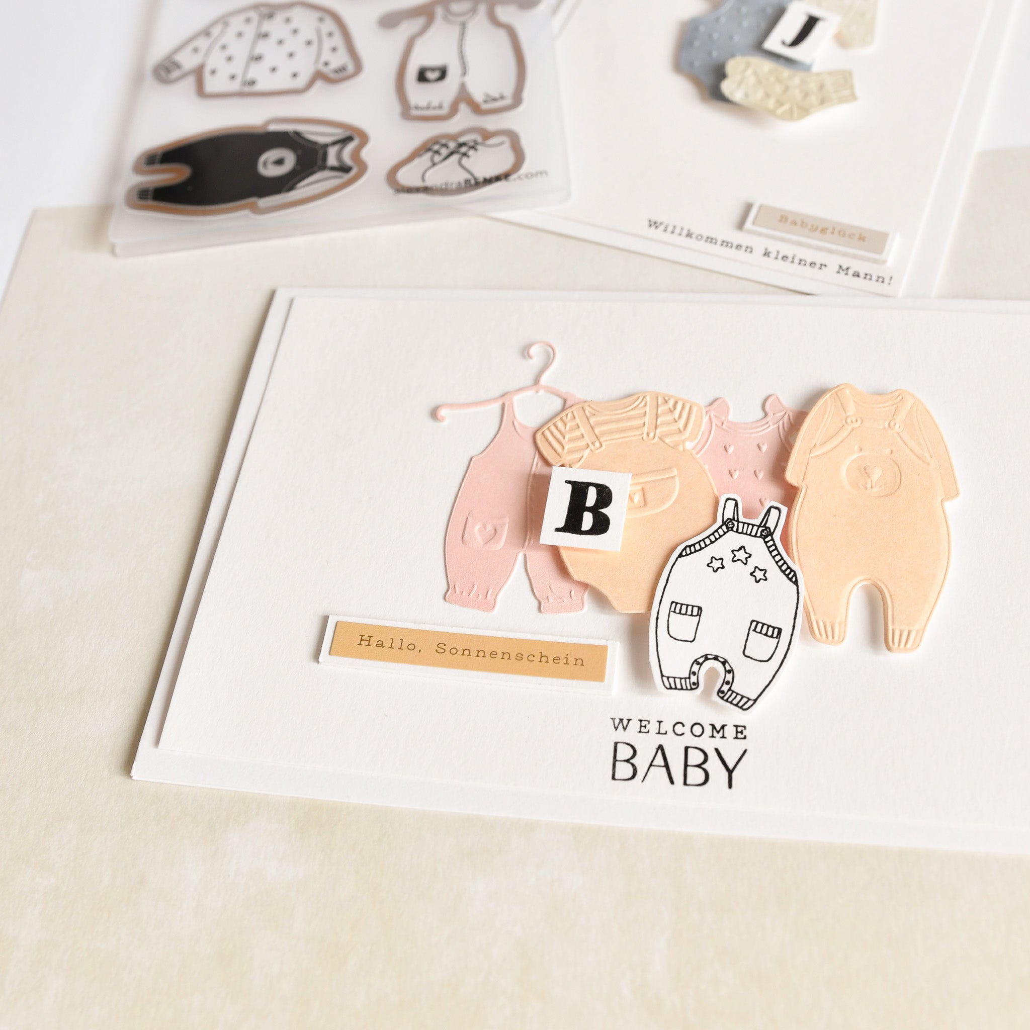 Grußkarte mit Alexandra Renke Designpapieren, ausgeschnittener Babykleidung, einem Block mit dem Buchstaben B und dem Text "Welcome Baby" auf hellem Hintergrund - wunderschön gestaltet mit unserem Cut & Emboss Folder.