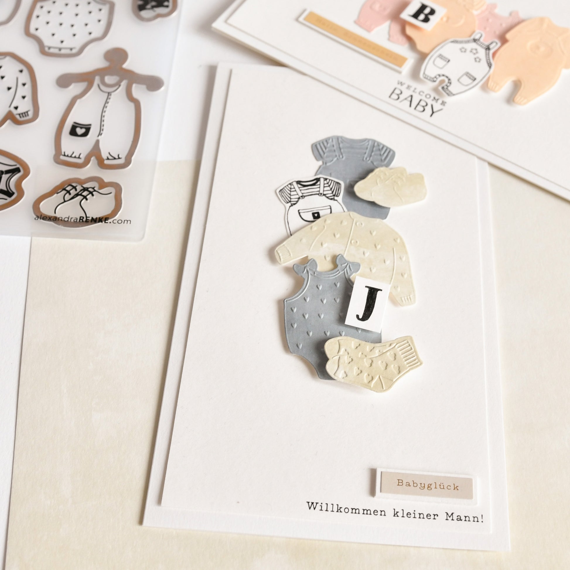 Diese Grußkarte von Alexandra Renke besteht aus hochwertigem Designpapier und einem Cut & Emboss Folder für ausgeschnittene Babykleidung und den Buchstaben J; der deutsche Text heißt einen kleinen Jungen herzlich willkommen.