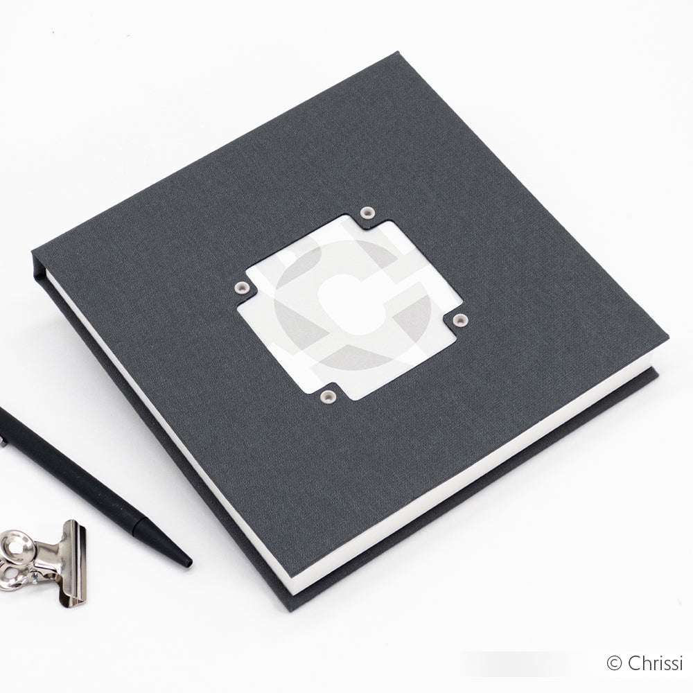 Quadratisches graues Notizbuch von Alexandra Renke mit einem ausgeschnittenen Fenster auf dem Einband, das mit Buchbindetechniken und präzisem PAPPcut hergestellt wurde, neben einem Stift und einem kleinen Metallclip.