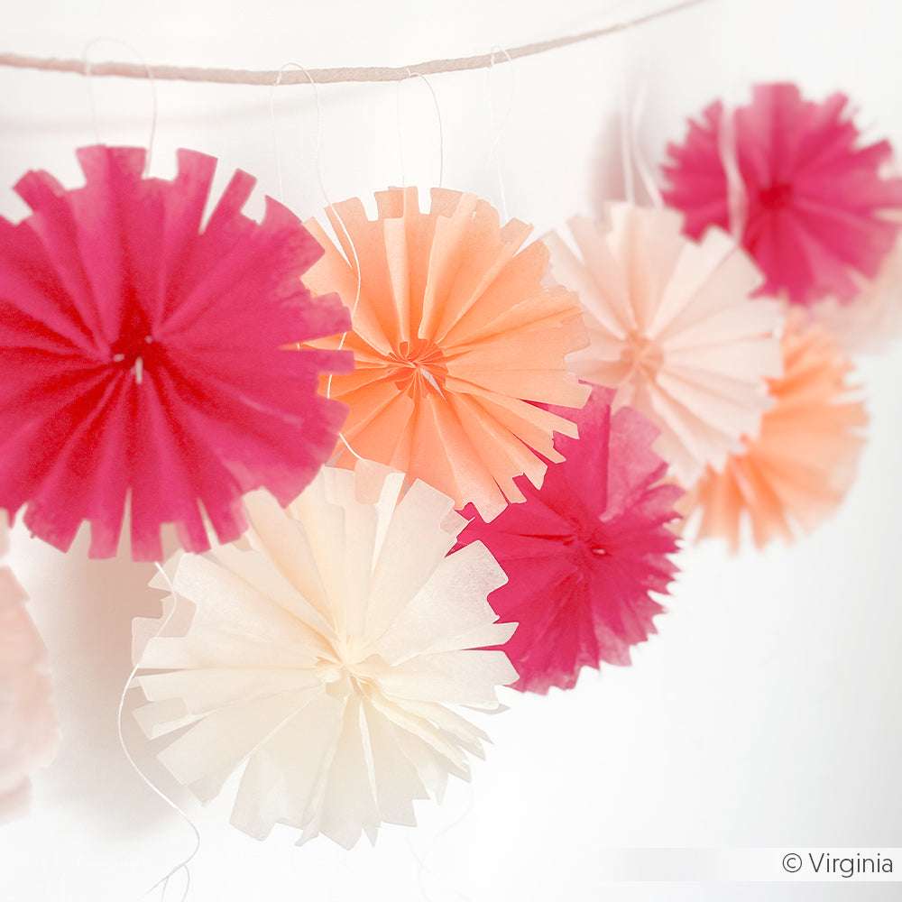 Die rosafarbenen, pfirsichfarbenen und weißen Papierrosetten von Alexandra Renke, die mit Stahlbandstanzen und PAPPcut-Werkzeugen hergestellt wurden, hängen als festliche Dekoration vor einem weißen Hintergrund.