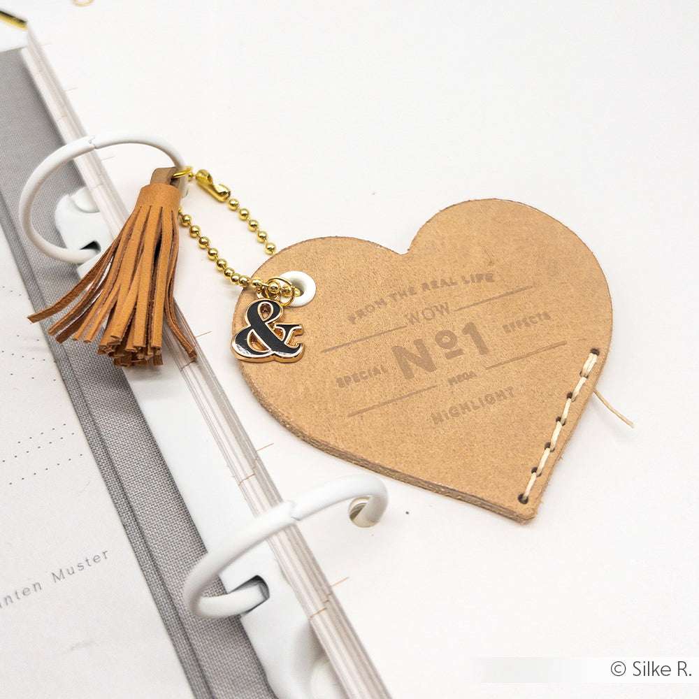 Herzförmiges Lederlesezeichen mit Quaste und Anhängern aus dem Glyphen Trio von Alexandra Renke auf einem gerahmten Planerblatt.