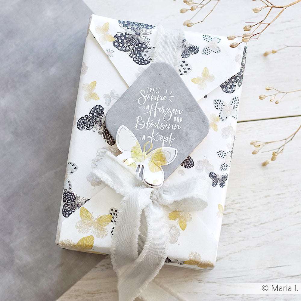 Geschenkverpackung aus Papier mit Schmetterlingsmuster von Alexandra Renke mit weißem Band und einem PAPPcut-Schmetterlingsanhänger auf einer hellen Holzunterlage.