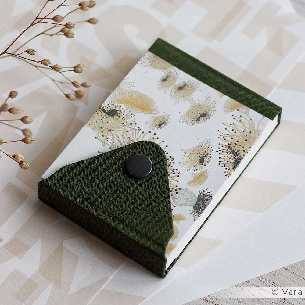 Auf einer gemusterten Oberfläche liegt ein in Buchbindetechnik gefertigtes, floral gemustertes Notizbuch von Alexandra Renke mit grünem Stoffeinband und Druckknopf.