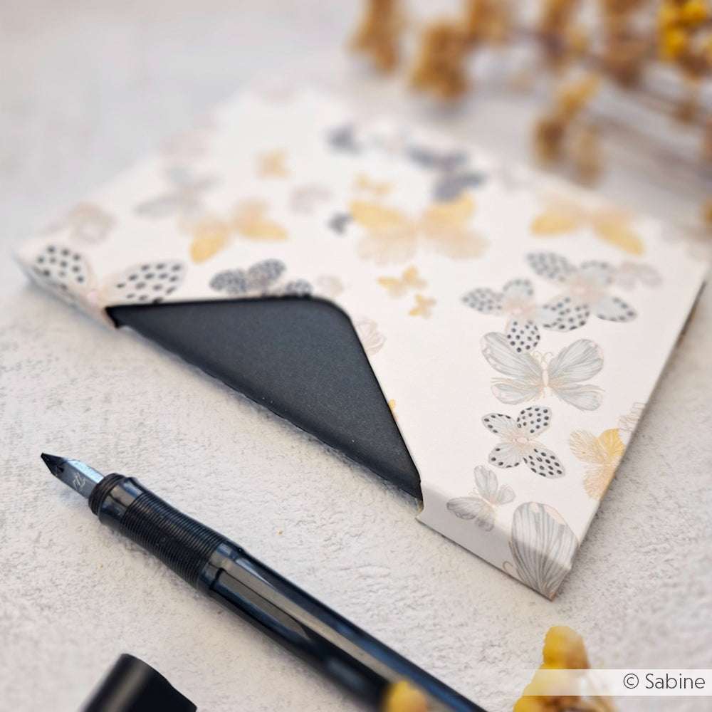 Ein Füllfederhalter neben einem Umschlag mit Blumenmuster und einer schwarzen Karte auf einer hellen Fläche - ideal, um Alexandra Renke-Produkte vorzustellen oder Ihre Lieblings-Papierbasteltechniken hervorzuheben.