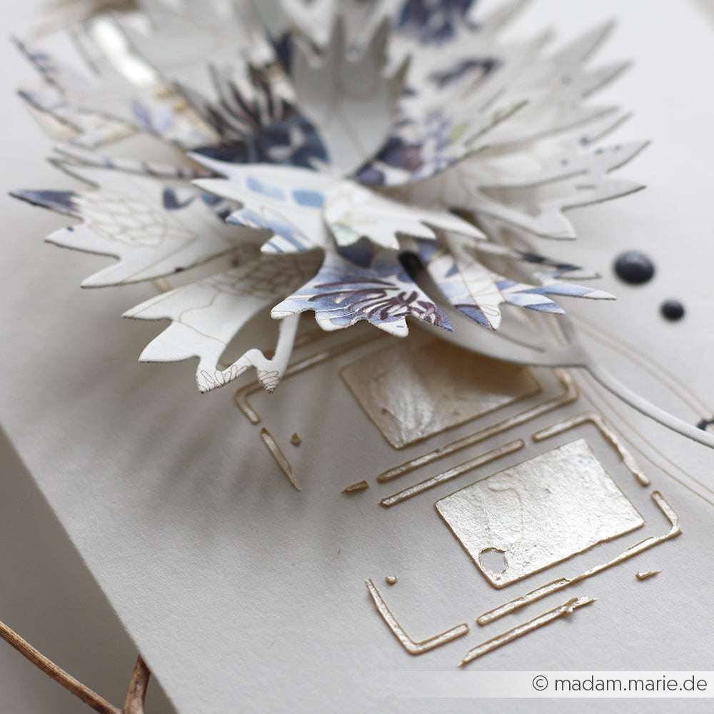 Nahaufnahme einer handgefertigten Karte von Alexandra Renke mit geschichteten Papierblättern und metallisch geprägten geometrischen Formen, die mit Schablonen für zusätzliche Textur und Details hergestellt wurden.